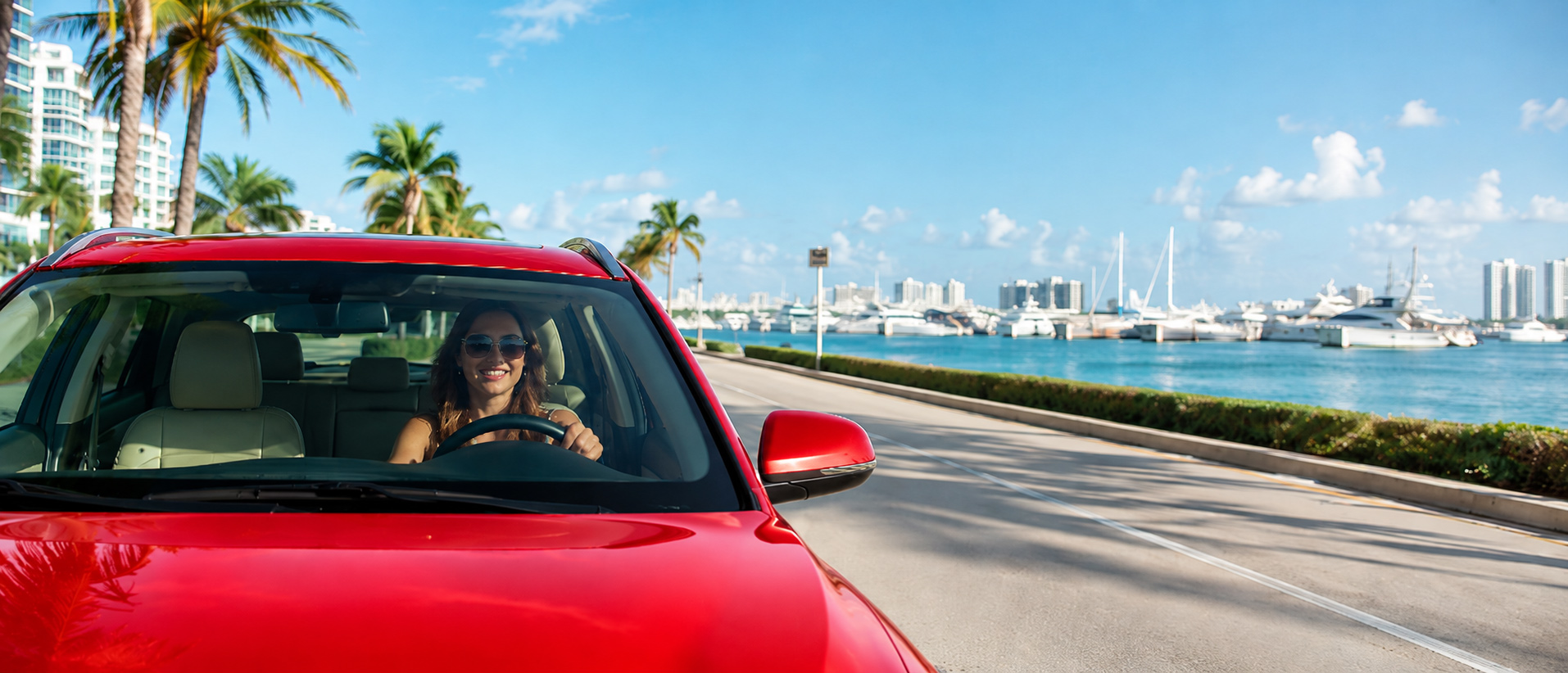 Happy woman driving a red car on a coastal road beside a Miami-style marina with boats and ocean views